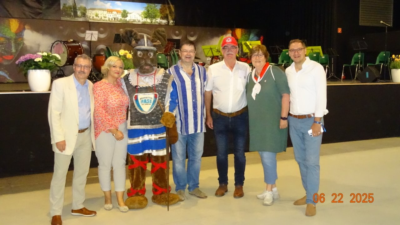 Sechs Personen und eine Person in bayerischem Trachtenkostüm mit Hut, Maske und Stock stehen in einer Halle vor einer Bühne mit Musikinstrumenten.