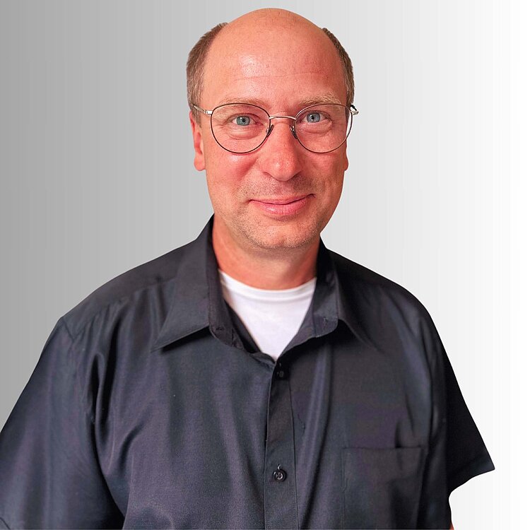 Mann mit Brille lächelt in die Kamera. Er trägt ein dunkles Hemd. Hintergrund ist hellgrau.