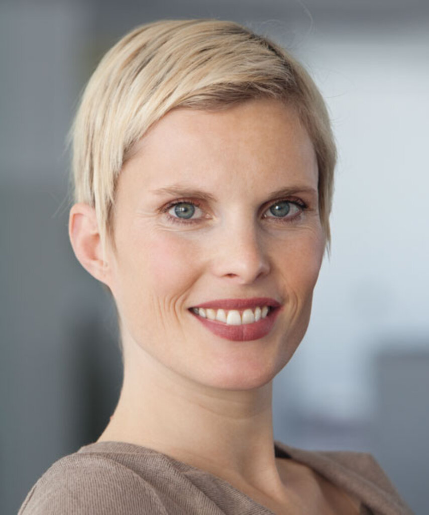 Person mit kurzen blonden Haaren trägt braunen Pullover und sitzt vor unscharfem Hintergrund