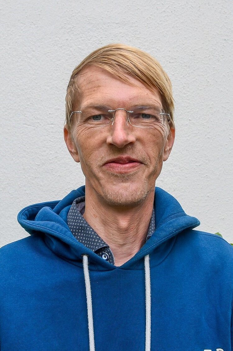 Person mit Brille und blauem Hoodie lächelt vor neutralem Hintergrund.