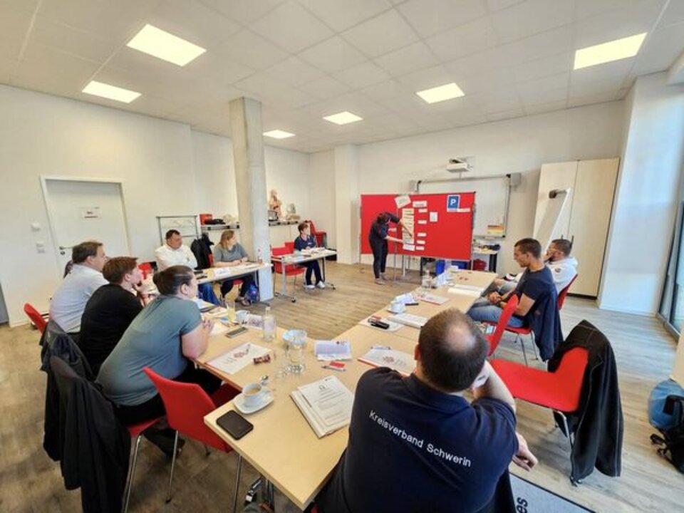 Menschen sitzen an Tischen in einem Seminarraum, ein Redner zeigt auf eine Tafel.