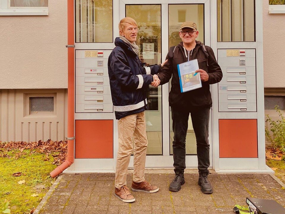 Zwei Männer schütteln sich vor einem Gebäude die Hand. Einer hält eine Mappe, im Hintergrund ist ein Briefkasten.