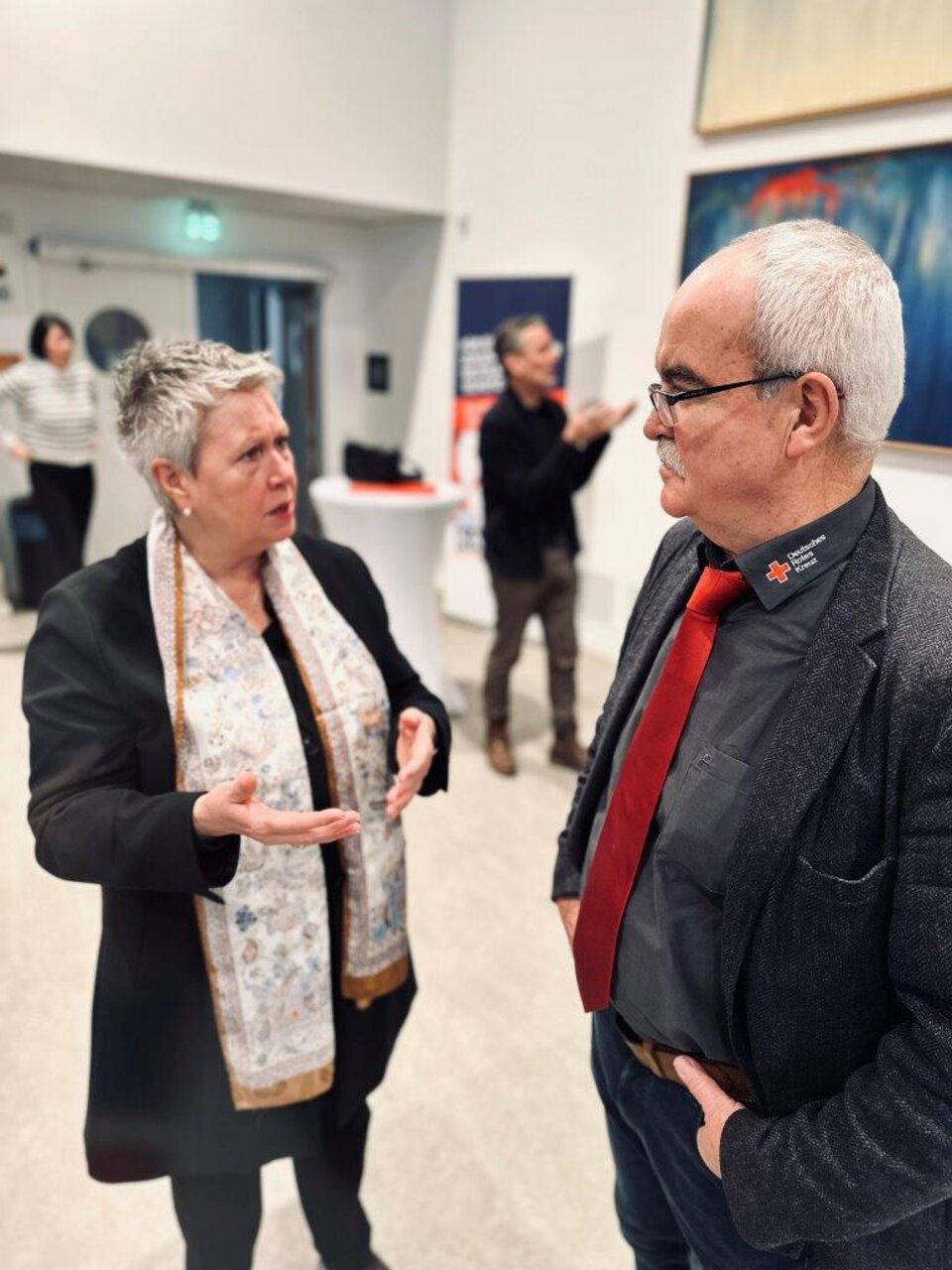 Zwei Personen diskutieren in einem Raum, dahinter weitere Personen und ein Stehtisch.