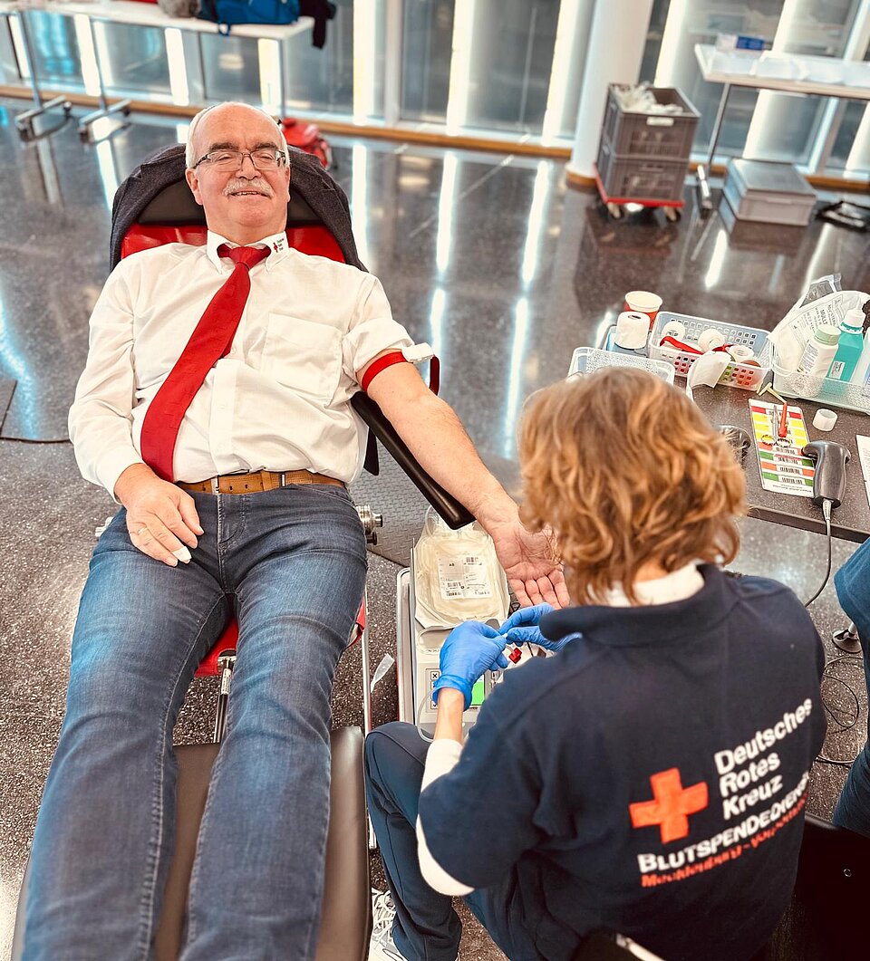 Mann spendet Blut, liegt auf Liege; DRK-Mitarbeiterin entnimmt Blut, umgeben von medizinischer Ausrüstung.