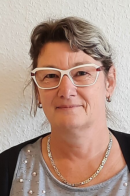 Person mit Brille vor einer hellen Wand, trägt eine Kette und ein graues Oberteil mit Perlendetails.