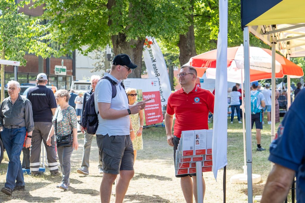 Messe mit Ständen unter Bäumen, Personen in Gesprächen, rot-weißer Sonnenschirm, Banner im Hintergrund.