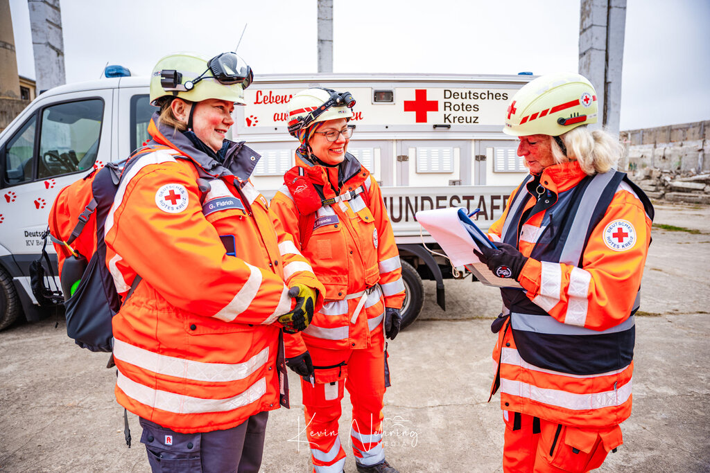 Drei Personen in Rettungskleidung vor DRK-Fahrzeug, eine liest von einem Klemmbrett.